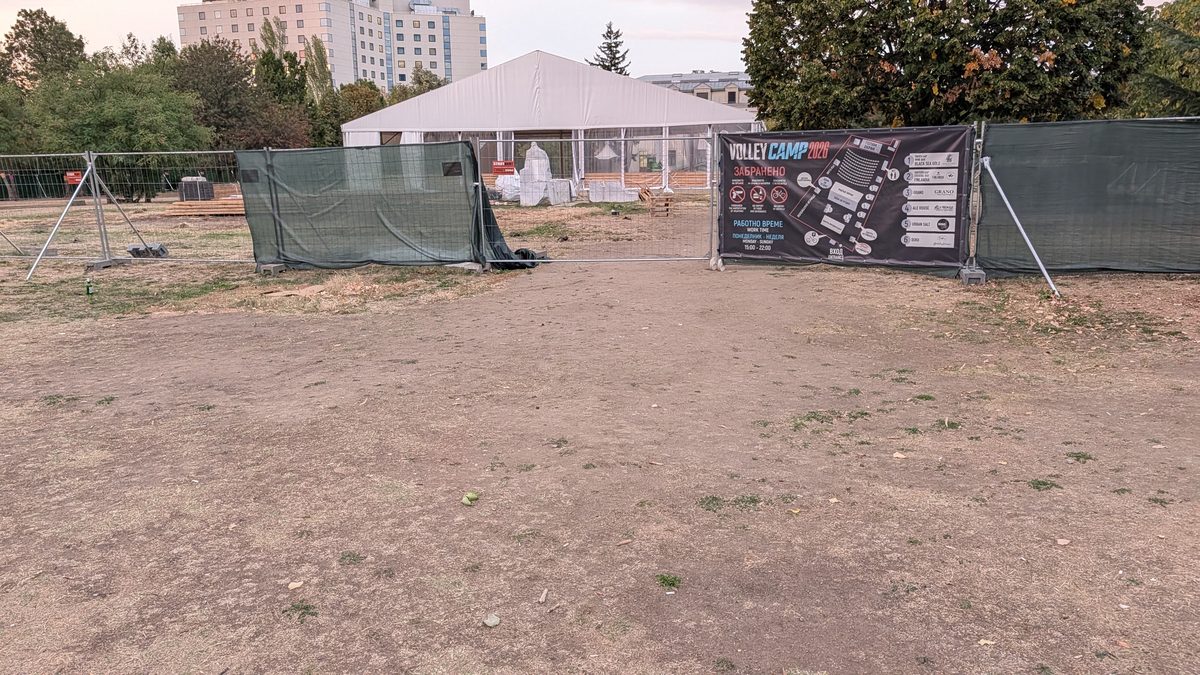 Поляната в Южен парк II на 29 септември 2026 г. Метална ограда със зелена мрежа и банер
                      „Volley Camp 2026“ с карта на зоната и списък със заведения. Зад оградата стоят голяма
                      бяла шатра, палети и опаковани декори. На преден план - гола пръст без трева.
